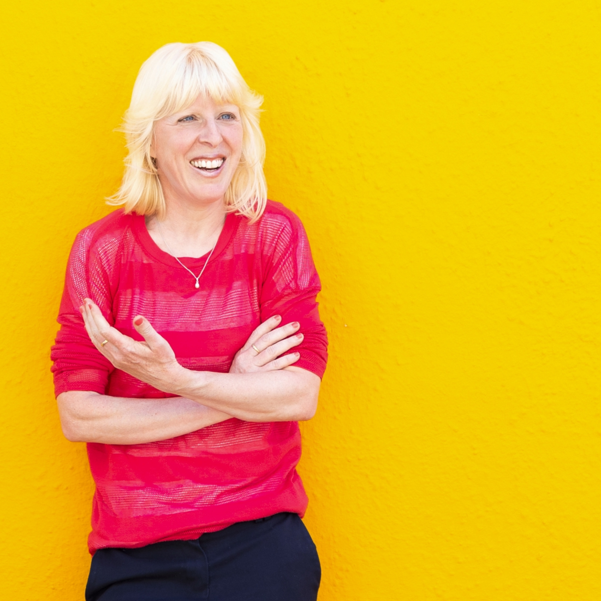 Leigh (a white female) leant against a mustard yellow wall. She's in a pink top with the sleeves rolled up, and navy trousers. She's smiling, and raising her left arm in a gesture.